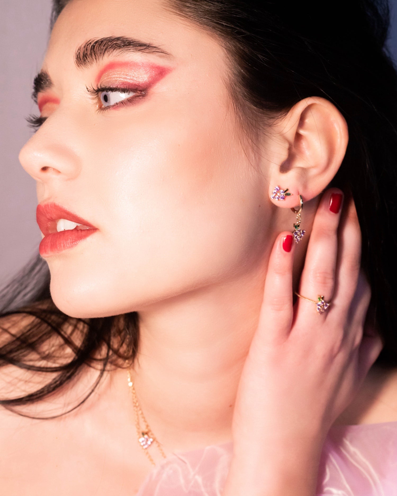 Primo piano di orecchini a lobo uva dorati Fruits & Jewels indossati con makeup elegante e manicure rossa