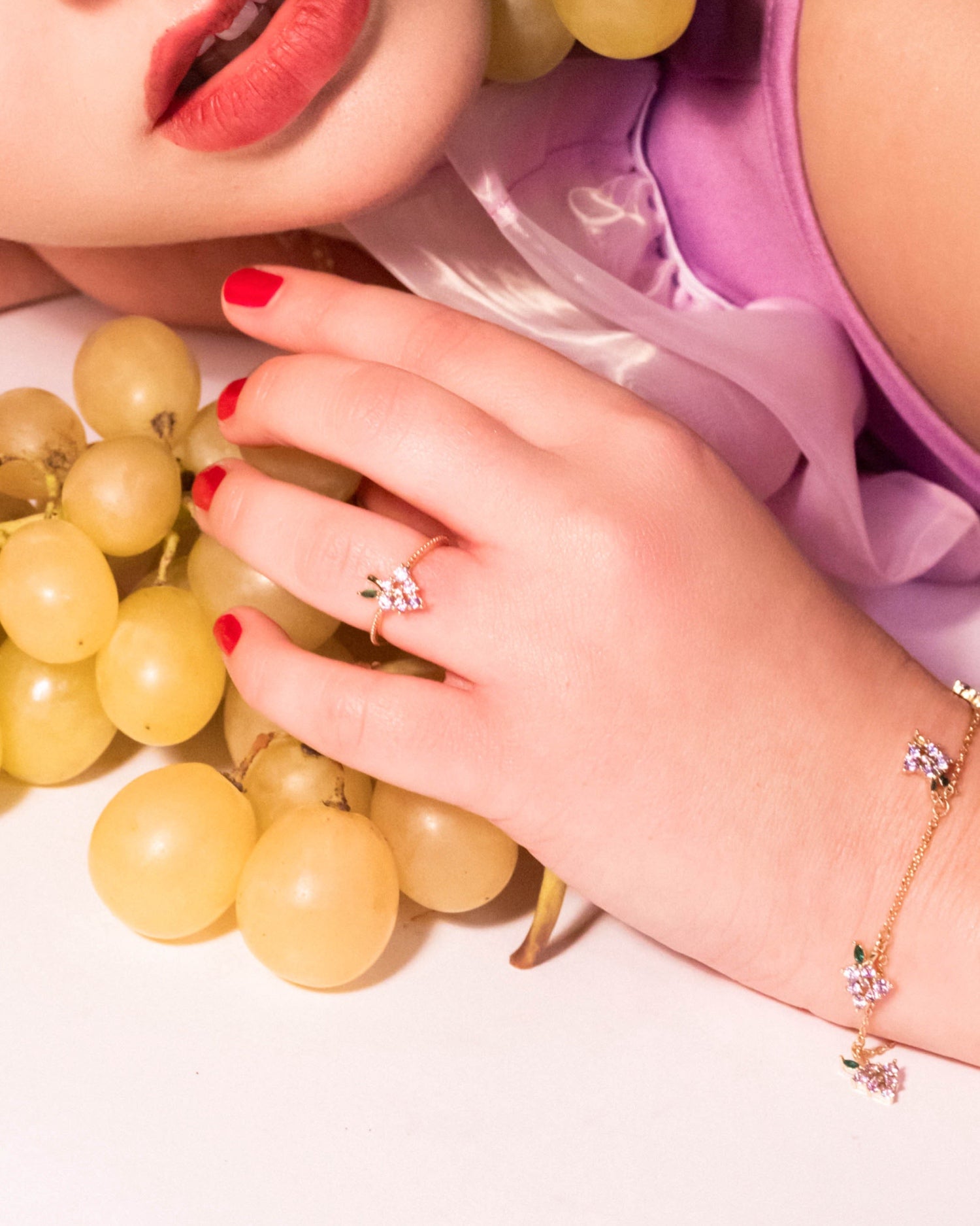 Mano con bracciale dorato con charms a grappolo d’uva e anello coordinato su sfondo bianco con chicchi d’uva gialla