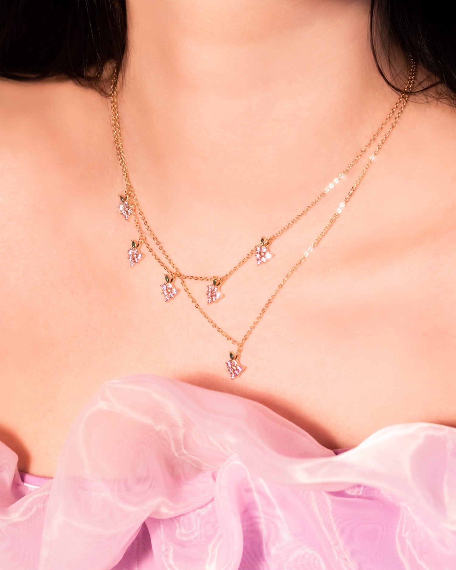 Primo piano su una collana placcata oro con cinque charms a forma di grappolo d’uva firmata Fruits & Jewels, indossata sopra un abito lilla in organza.