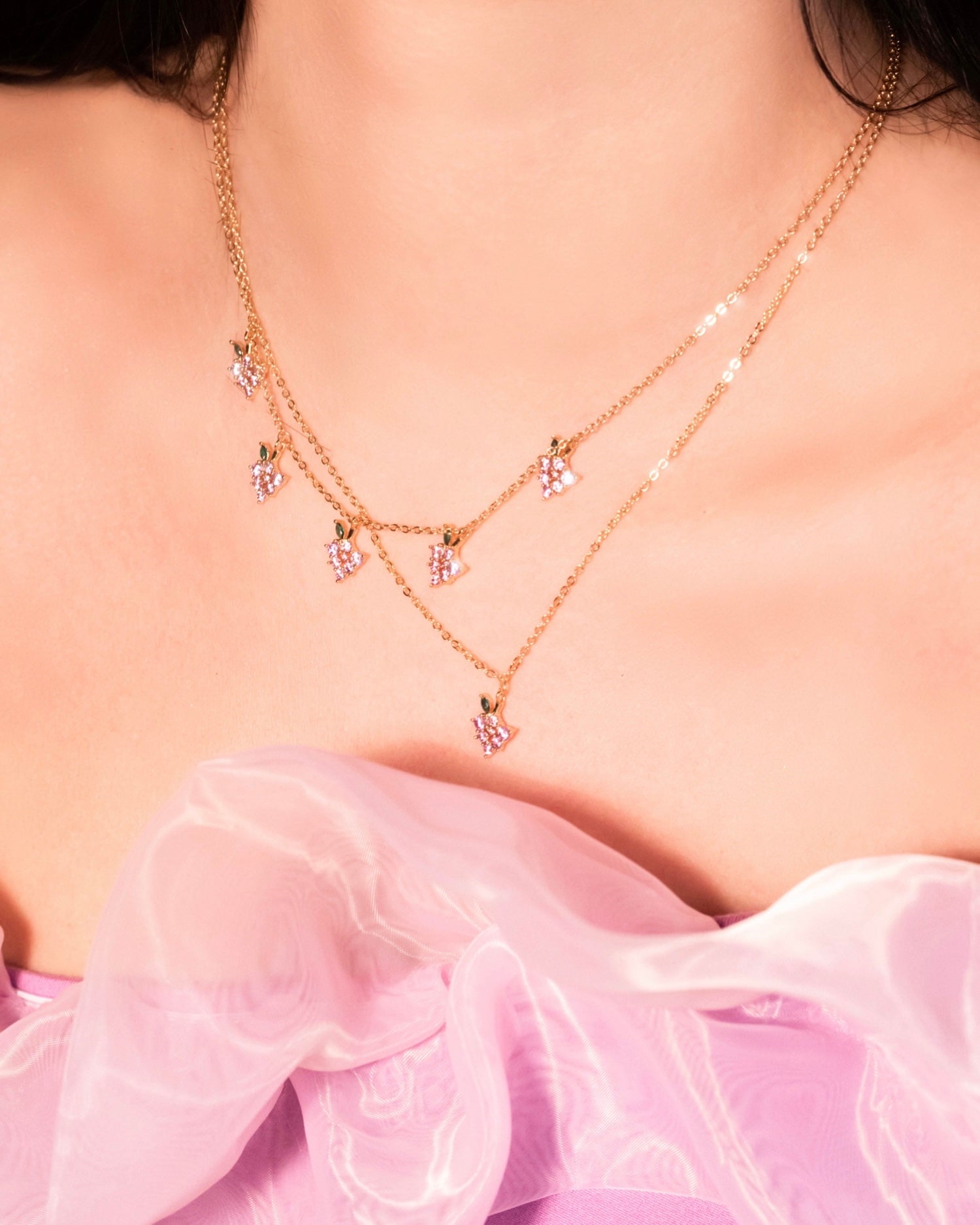 Décolleté con doppia collana dorata: una con ciondolo singolo a grappolo d’uva e l’altra con cinque charms a grappolo d’uva, indossate su un abito lilla in organza.
