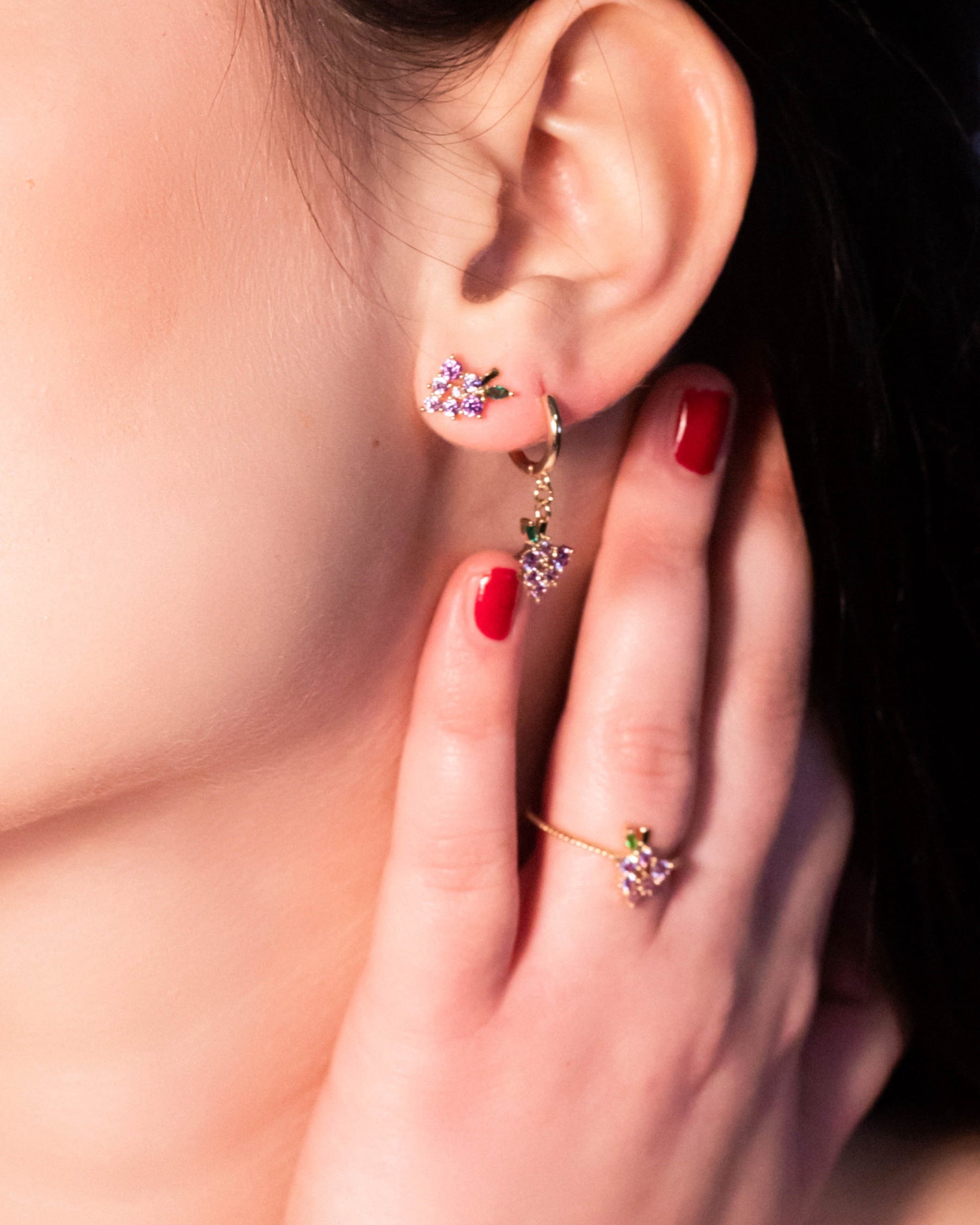 Dettaglio di orecchino uva dorato Fruits & Jewels con mano a evidenziare anello coordinato e manicure rossa