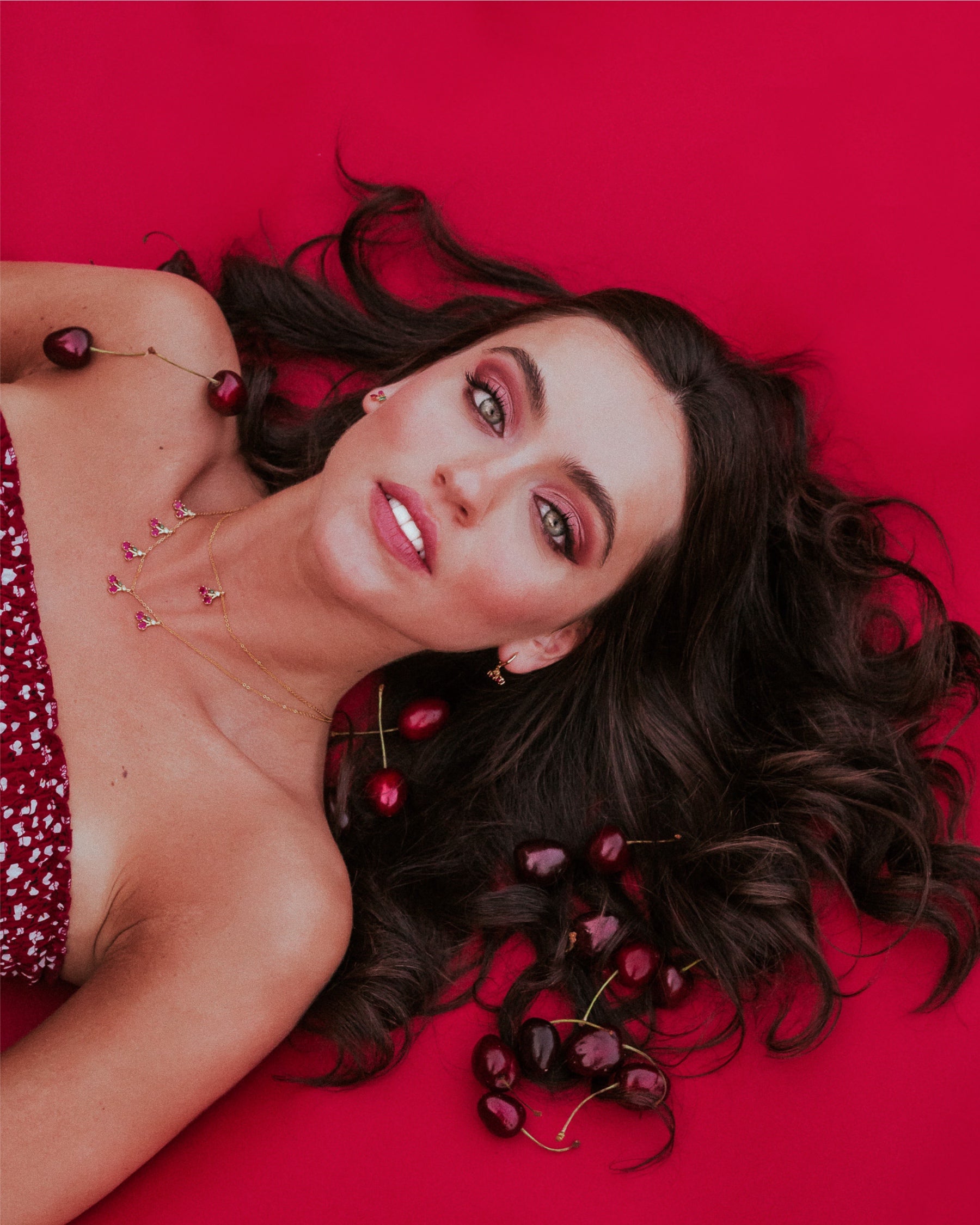 Modella distesa su sfondo rosso con collana charms ciliegia in oro 18k – gioiello fruttato e audace di Fruits & Jewels