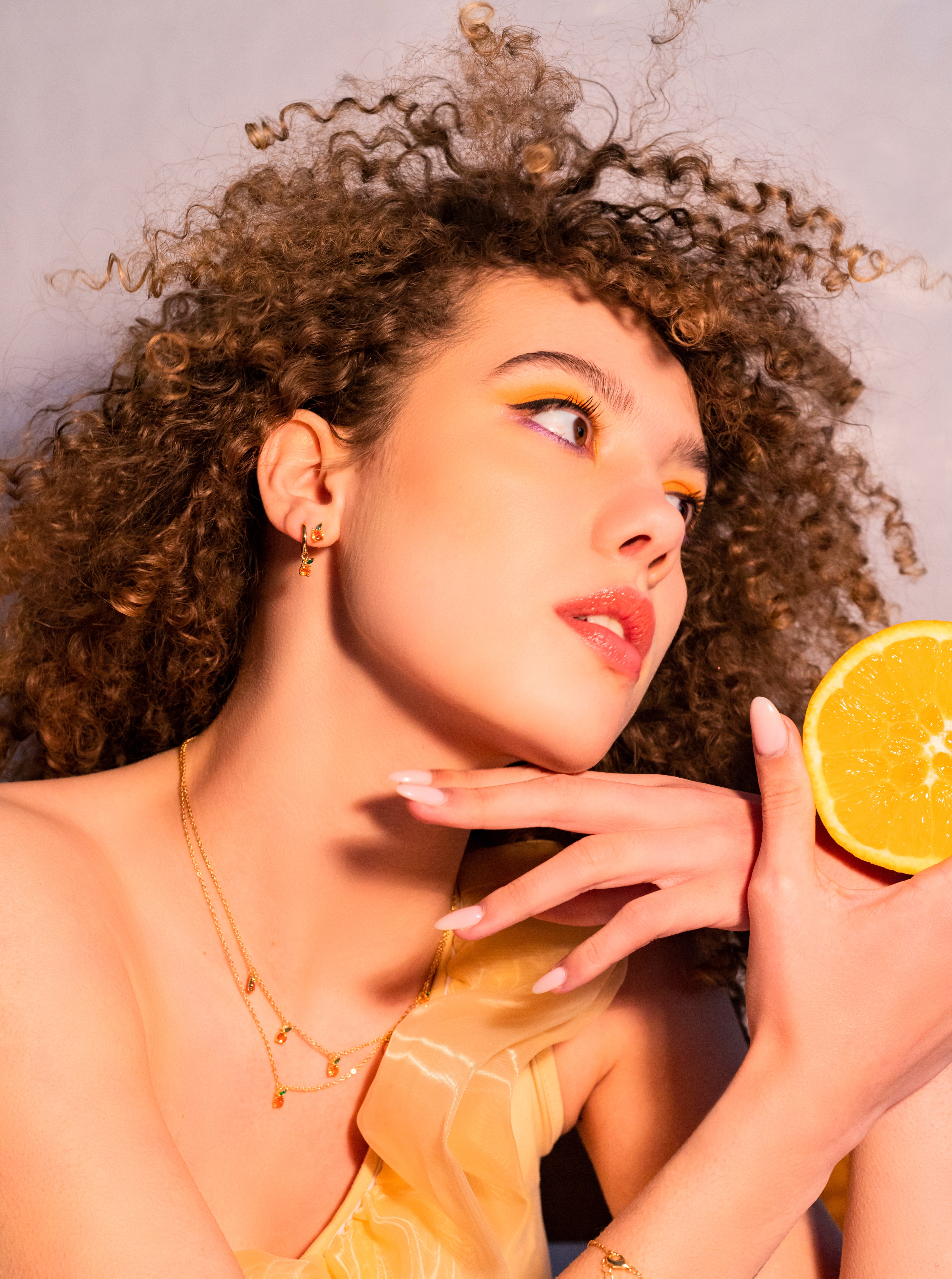Collana pendente arancia placcata oro 18 carati Fruits & Jewels indossata da modella riccia con sfondo neutro e fetta d’arancia