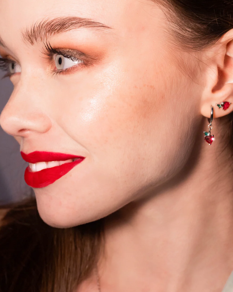 Orecchino pendente a forma di mela rossa, con finitura placcata in argento, indossato da modella con rossetto rosso.