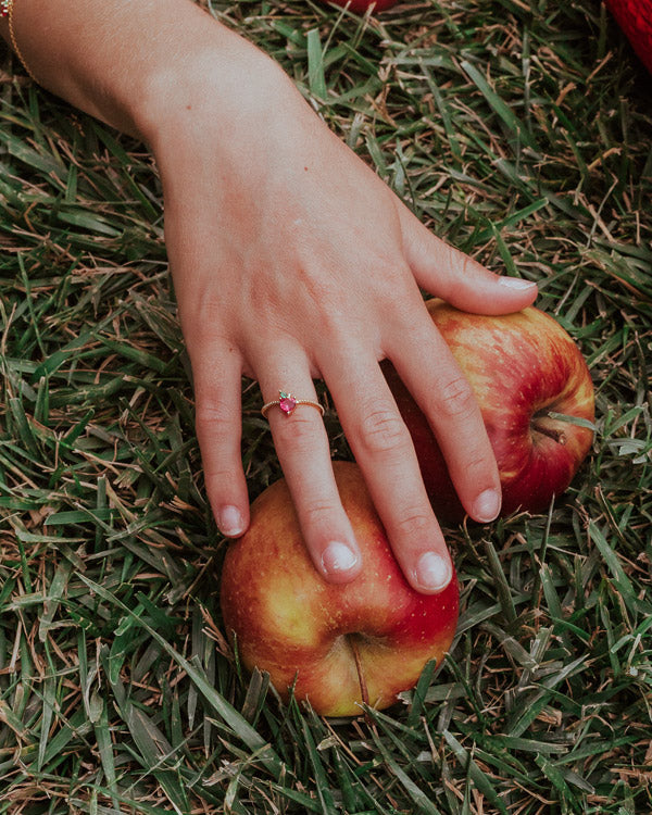 Dettaglio mano con anello mela appoggiata sull’erba vicino a due mele, Fruits & Jewels