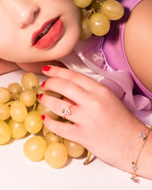 Anello uva placcato oro 18 carati Fruits & Jewels indossato su mano femminile con manicure rossa e grappolo d’uva gialla
