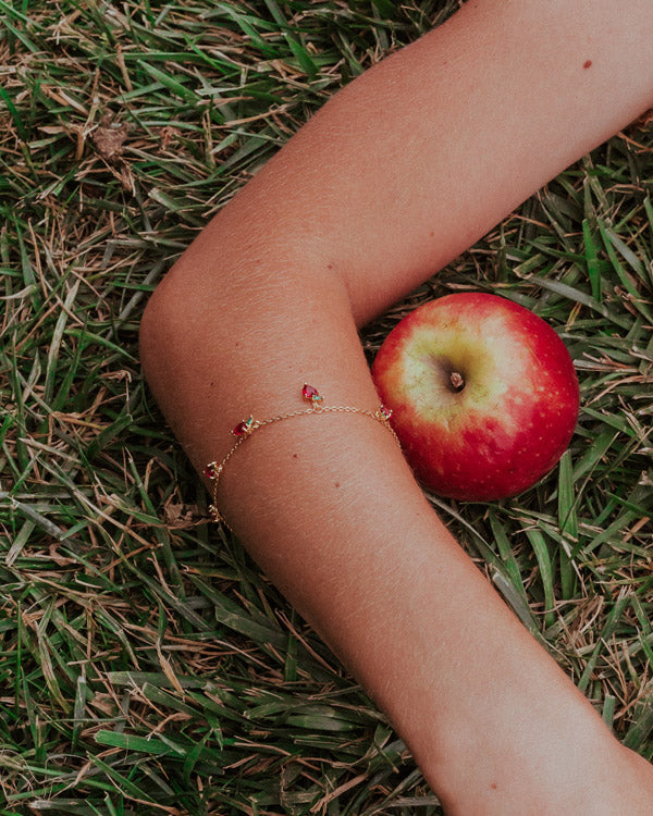 Dettaglio bracciale mela su polso disteso sull’erba vicino a una mela, Fruits & Jewels