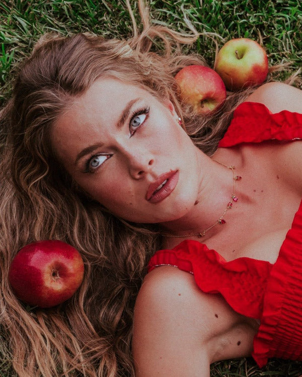 Ritratto artistico con collana charms mela placcata in oro 18 carati indossata su abito rosso, ambientazione con mele, Fruits & Jewels